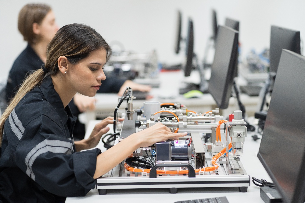 A woman works on a piece of machinery at a workstation with computer monitors. Other individuals are visible in the background, also focused on similar tasks.