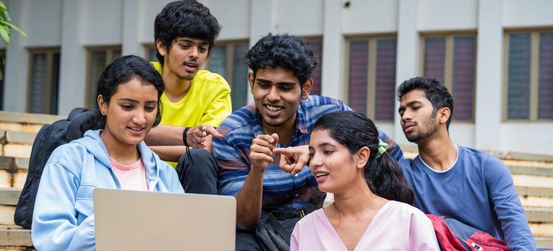 A group of five young adults sit on outdoor steps, looking at a laptop and discussing something.