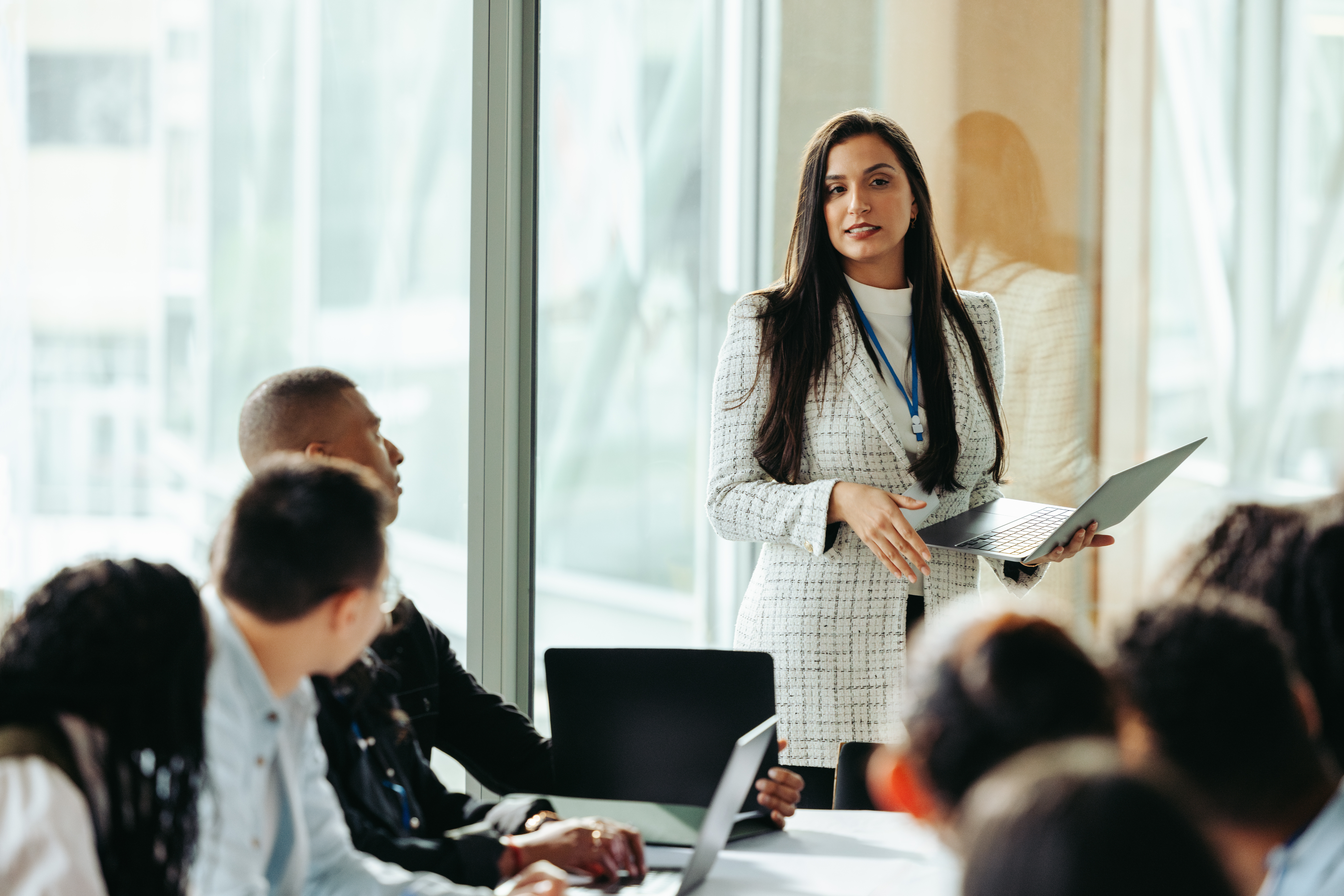 Confident female business owner leading a board meeting, using a laptop to present strategies to diverse team in a modern office setting