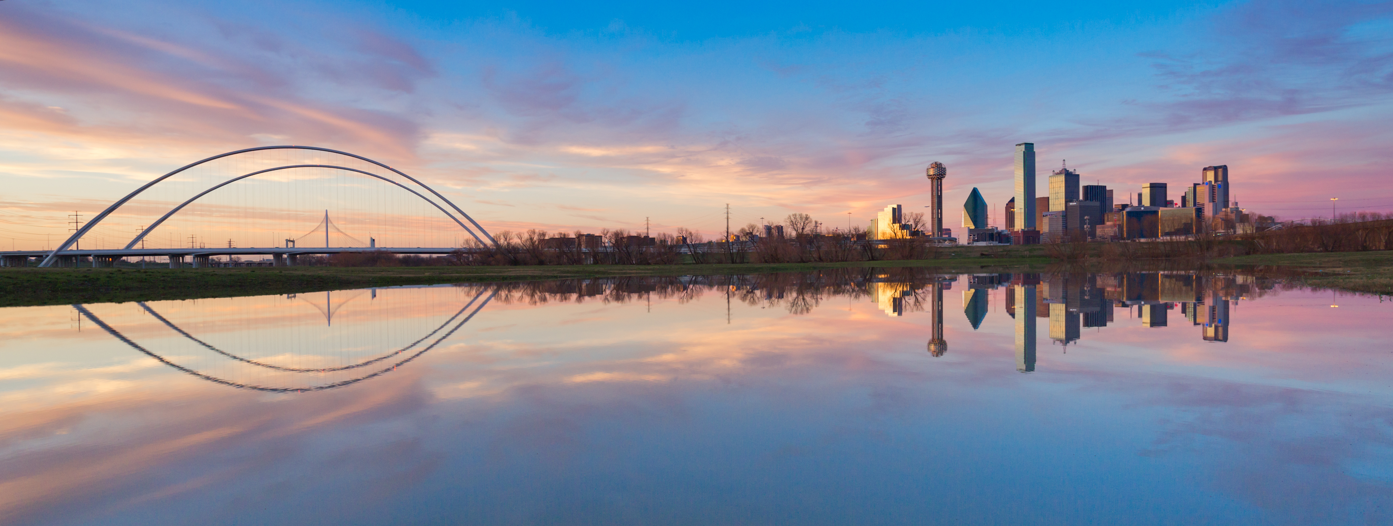 Dallas skyline and Margaret Hunt Hill Bridge reflected in calm water at sunrise with colorful clouds in the sky.