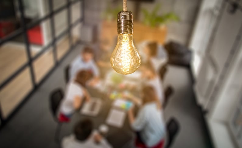 An illuminated light bulb is in focus in the foreground, with four people working around a table in an office setting in the background.