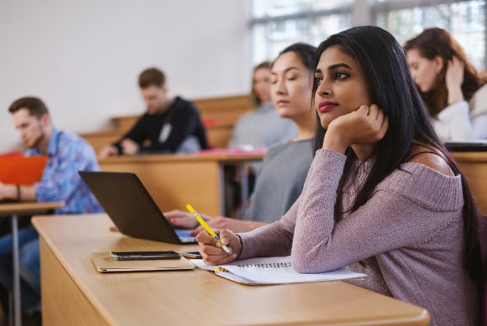 A diverse group of students focused on their studies in a classroom, with a young south asian woman in the foreground looking thoughtful.