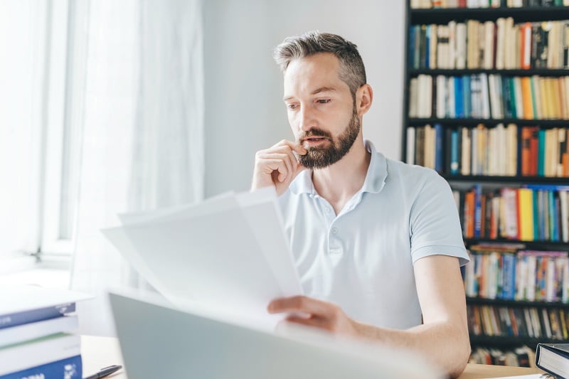 A recent graduate with a beard and short hair sits at a desk in front of a bookshelf, holding documents and looking thoughtful.