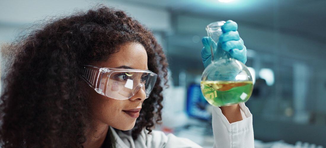 A scientist wearing protective eyewear and gloves examines a conical flask containing a yellow-green liquid in a laboratory setting.