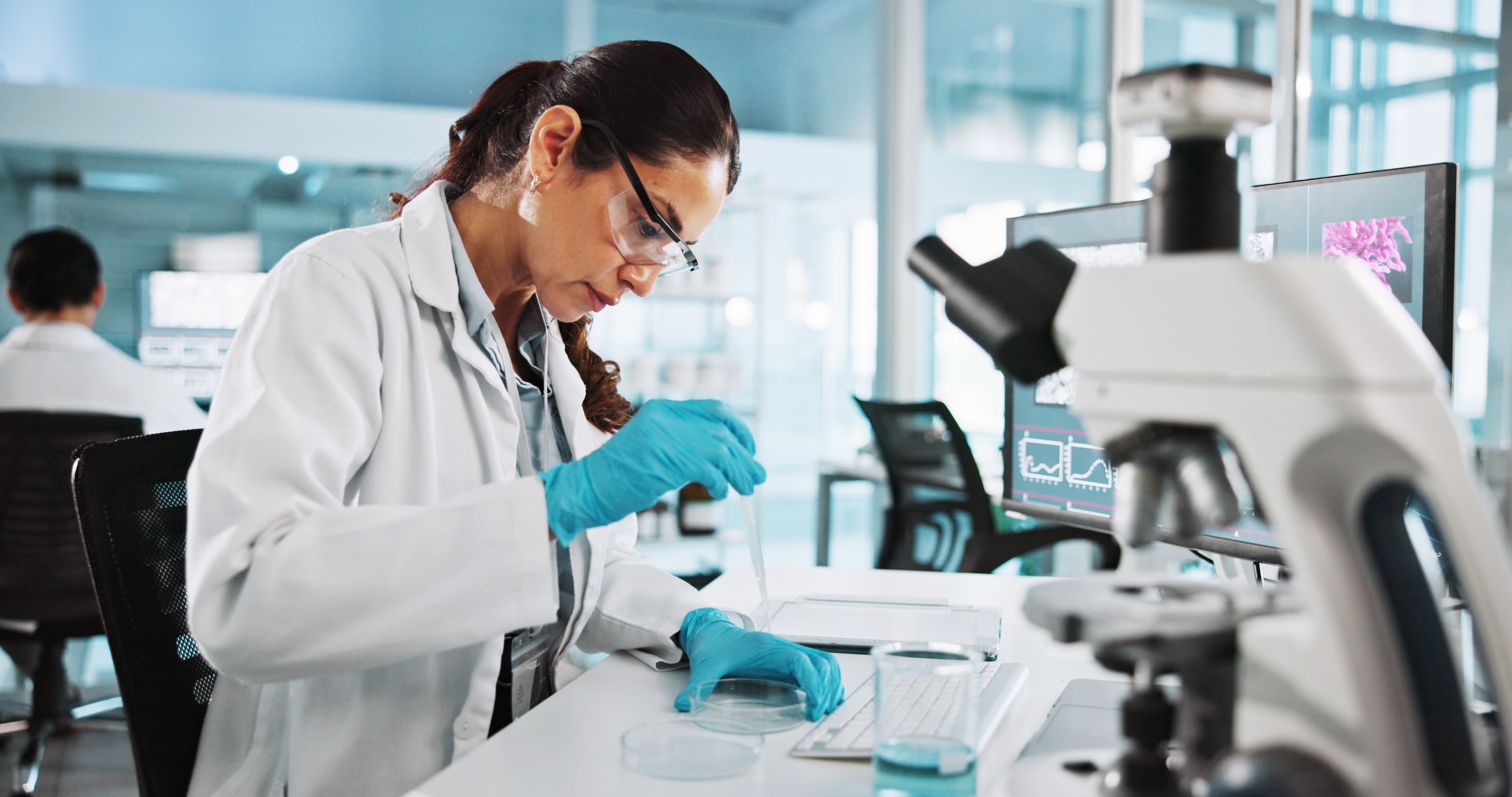 Female scientist in a modern lab wearing protective gear, performing an experiment with a pipette and petri dishes, with a microscope and computer monitors nearby.
