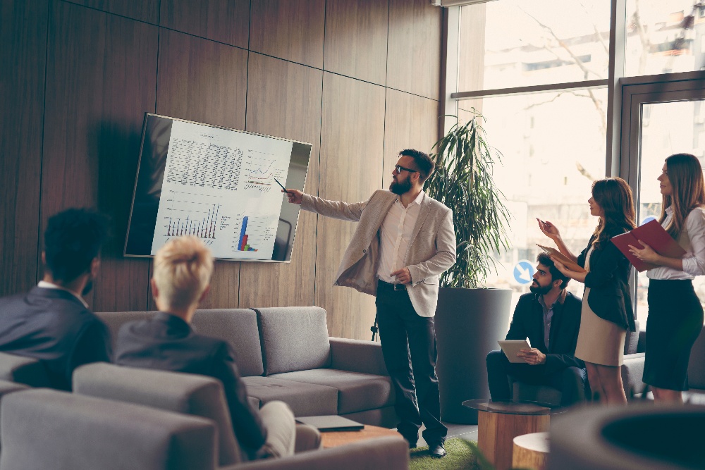 A man in formal attire is presenting data on a wall-mounted screen to a small group of people seated on couches in a modern office setting.