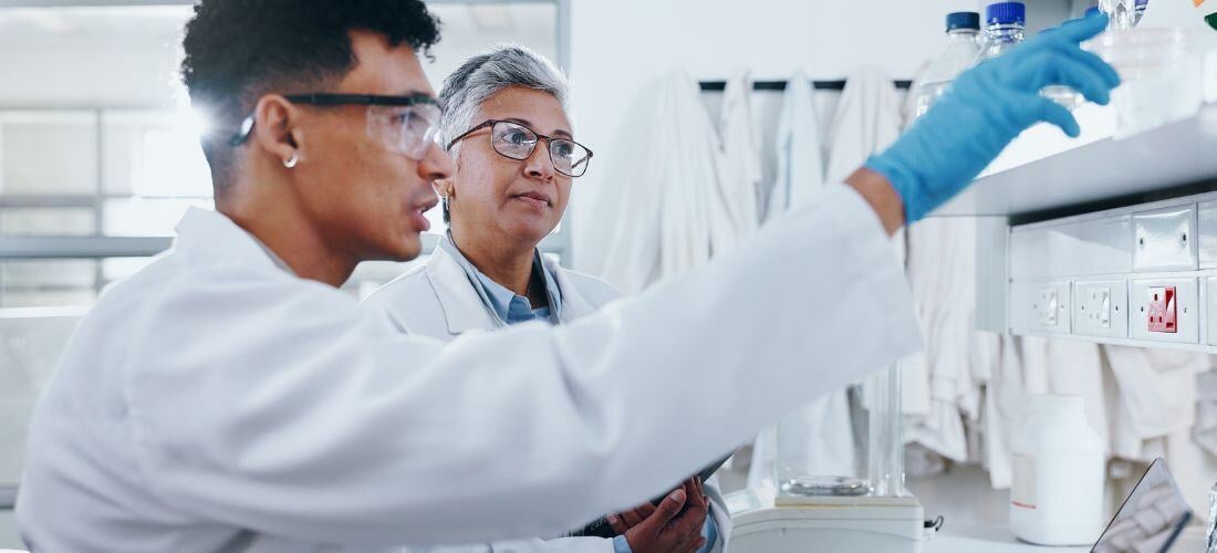 Two scientists in lab coats and safety glasses work in a laboratory. One points at a shelf with lab equipment, while the other observes.