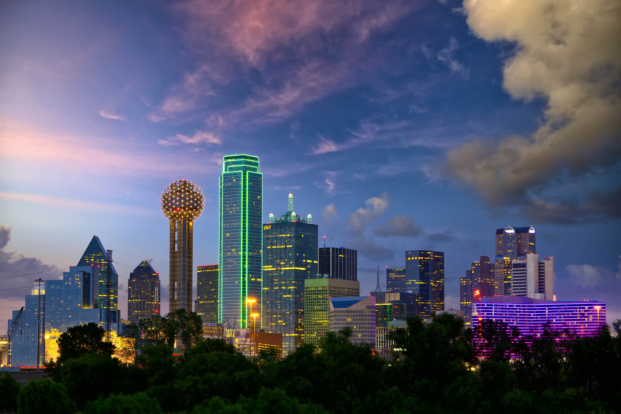 Dallas skyline at twilight with vibrant clouds, featuring distinctive reunion tower and modern skyscrapers illuminated against the dusk sky.